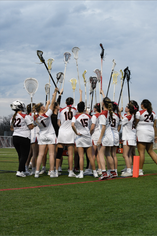 A girls lacrosse team in white uniforms huddles on a turf field with their sticks raised high in the air under a cloudy sky.