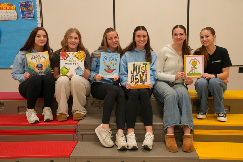Six students sit on colorful tiered seating in a classroom, smiling and holding up brightly illustrated children’s books. A bulletin board is visible in the background, creating a welcoming, literacy-focused setting.