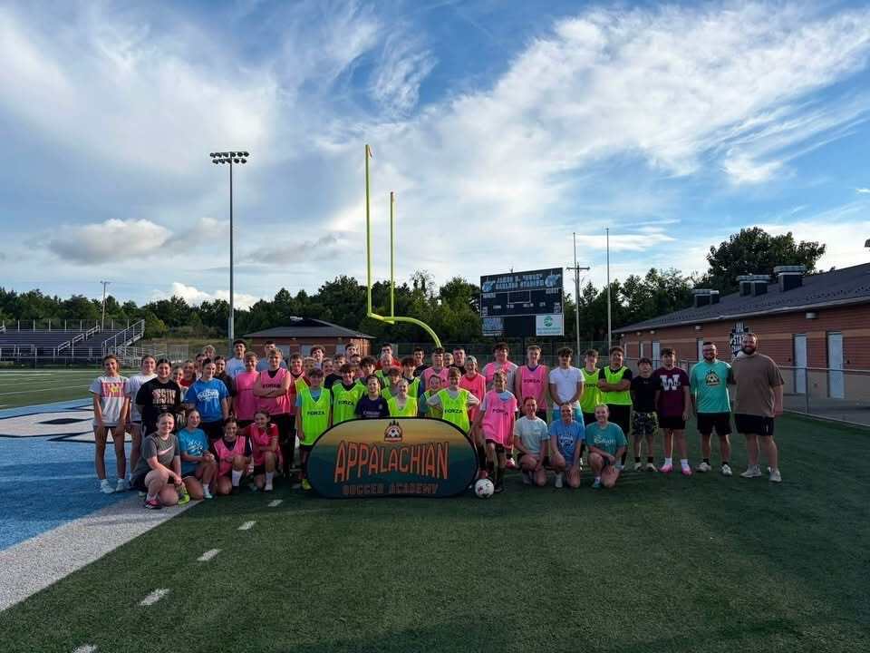 Photo 1: A group of youth soccer players and coaches pose behind an Appalachian Soccer Academy banner on Mingo Central's football field under a bright summer sky during the free soccer clinic.  Photo 2: Coach Noah Rhodes addresses a group of soccer campers gathered in a circle at midfield while participants stand with soccer balls on Mingo Central's turf field.  Photo 3: Camp participants run through soccer drills across the football field during an evening practice session as clouds gather overhead.  Photo 4: A large group of soccer campers, coaches, and volunteers pose together behind the Appalachian Soccer Academy banner on Mingo Central's football field following the conclusion of the free community soccer clinic.