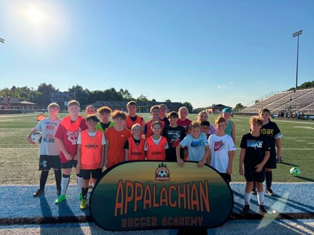 Photo 1: A group of youth soccer players and coaches pose behind an Appalachian Soccer Academy banner on Mingo Central's football field under a bright summer sky during the free soccer clinic.  Photo 2: Coach Noah Rhodes addresses a group of soccer campers gathered in a circle at midfield while participants stand with soccer balls on Mingo Central's turf field.  Photo 3: Camp participants run through soccer drills across the football field during an evening practice session as clouds gather overhead.  Photo 4: A large group of soccer campers, coaches, and volunteers pose together behind the Appalachian Soccer Academy banner on Mingo Central's football field following the conclusion of the free community soccer clinic.