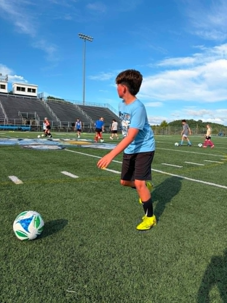Photo 1: A group of youth soccer players and coaches pose behind an Appalachian Soccer Academy banner on Mingo Central's football field under a bright summer sky during the free soccer clinic.  Photo 2: Coach Noah Rhodes addresses a group of soccer campers gathered in a circle at midfield while participants stand with soccer balls on Mingo Central's turf field.  Photo 3: Camp participants run through soccer drills across the football field during an evening practice session as clouds gather overhead.  Photo 4: A large group of soccer campers, coaches, and volunteers pose together behind the Appalachian Soccer Academy banner on Mingo Central's football field following the conclusion of the free community soccer clinic.