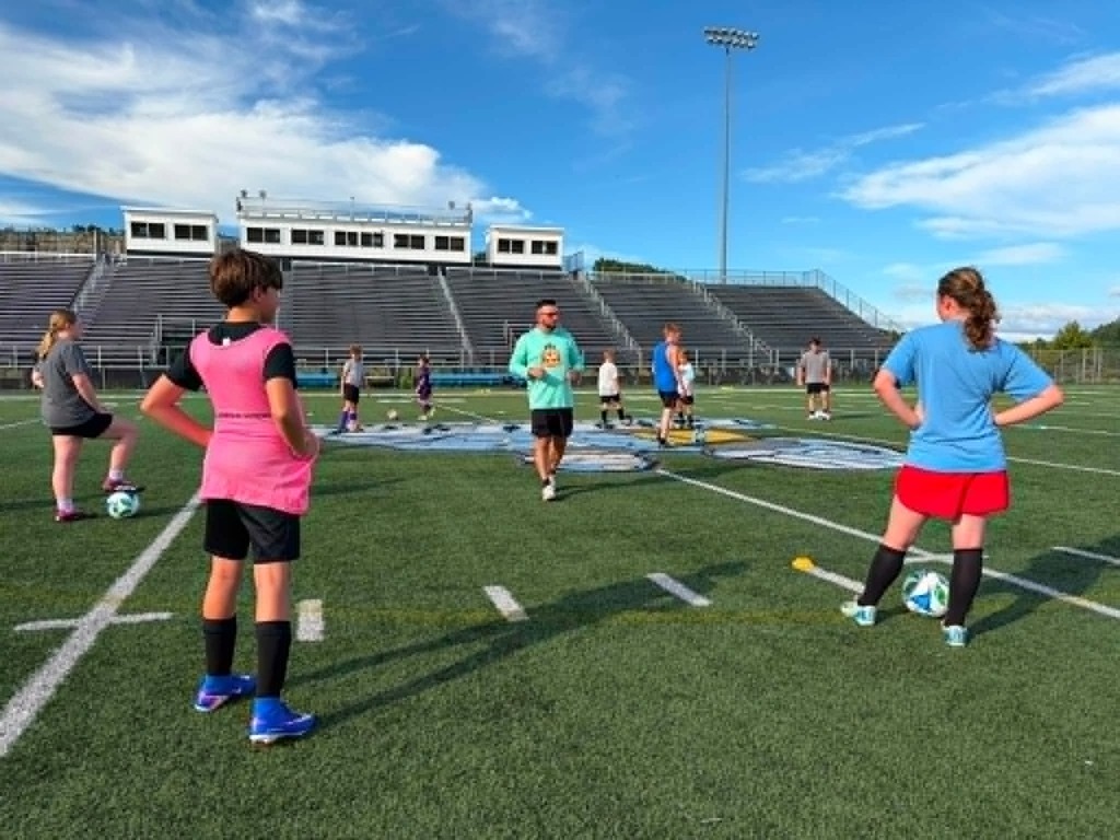 Photo 1: A group of youth soccer players and coaches pose behind an Appalachian Soccer Academy banner on Mingo Central's football field under a bright summer sky during the free soccer clinic.  Photo 2: Coach Noah Rhodes addresses a group of soccer campers gathered in a circle at midfield while participants stand with soccer balls on Mingo Central's turf field.  Photo 3: Camp participants run through soccer drills across the football field during an evening practice session as clouds gather overhead.  Photo 4: A large group of soccer campers, coaches, and volunteers pose together behind the Appalachian Soccer Academy banner on Mingo Central's football field following the conclusion of the free community soccer clinic.