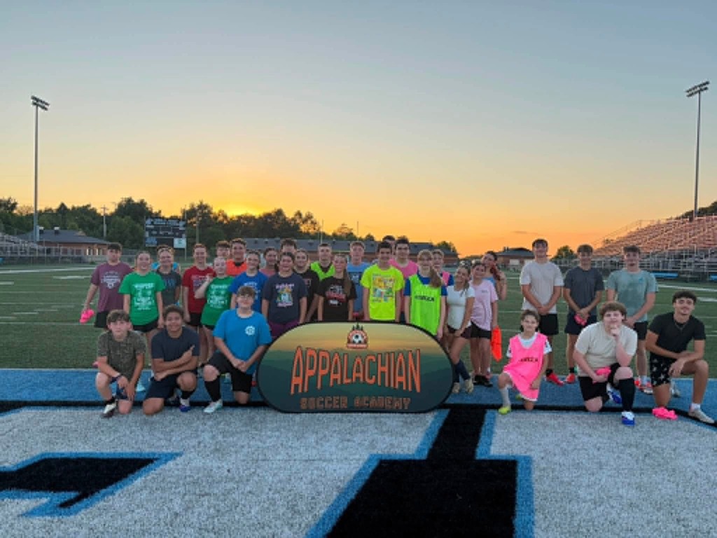 Photo 1: A group of youth soccer players and coaches pose behind an Appalachian Soccer Academy banner on Mingo Central's football field under a bright summer sky during the free soccer clinic.  Photo 2: Coach Noah Rhodes addresses a group of soccer campers gathered in a circle at midfield while participants stand with soccer balls on Mingo Central's turf field.  Photo 3: Camp participants run through soccer drills across the football field during an evening practice session as clouds gather overhead.  Photo 4: A large group of soccer campers, coaches, and volunteers pose together behind the Appalachian Soccer Academy banner on Mingo Central's football field following the conclusion of the free community soccer clinic.