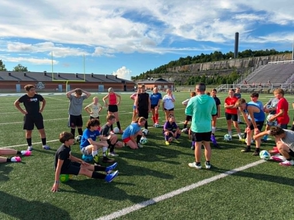 Photo 1: A group of youth soccer players and coaches pose behind an Appalachian Soccer Academy banner on Mingo Central's football field under a bright summer sky during the free soccer clinic.  Photo 2: Coach Noah Rhodes addresses a group of soccer campers gathered in a circle at midfield while participants stand with soccer balls on Mingo Central's turf field.  Photo 3: Camp participants run through soccer drills across the football field during an evening practice session as clouds gather overhead.  Photo 4: A large group of soccer campers, coaches, and volunteers pose together behind the Appalachian Soccer Academy banner on Mingo Central's football field following the conclusion of the free community soccer clinic.