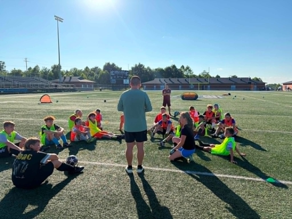 Photo 1: A group of youth soccer players and coaches pose behind an Appalachian Soccer Academy banner on Mingo Central's football field under a bright summer sky during the free soccer clinic.  Photo 2: Coach Noah Rhodes addresses a group of soccer campers gathered in a circle at midfield while participants stand with soccer balls on Mingo Central's turf field.  Photo 3: Camp participants run through soccer drills across the football field during an evening practice session as clouds gather overhead.  Photo 4: A large group of soccer campers, coaches, and volunteers pose together behind the Appalachian Soccer Academy banner on Mingo Central's football field following the conclusion of the free community soccer clinic.
