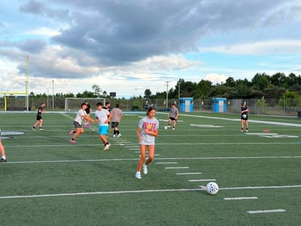 Photo 1: A group of youth soccer players and coaches pose behind an Appalachian Soccer Academy banner on Mingo Central's football field under a bright summer sky during the free soccer clinic.  Photo 2: Coach Noah Rhodes addresses a group of soccer campers gathered in a circle at midfield while participants stand with soccer balls on Mingo Central's turf field.  Photo 3: Camp participants run through soccer drills across the football field during an evening practice session as clouds gather overhead.  Photo 4: A large group of soccer campers, coaches, and volunteers pose together behind the Appalachian Soccer Academy banner on Mingo Central's football field following the conclusion of the free community soccer clinic.