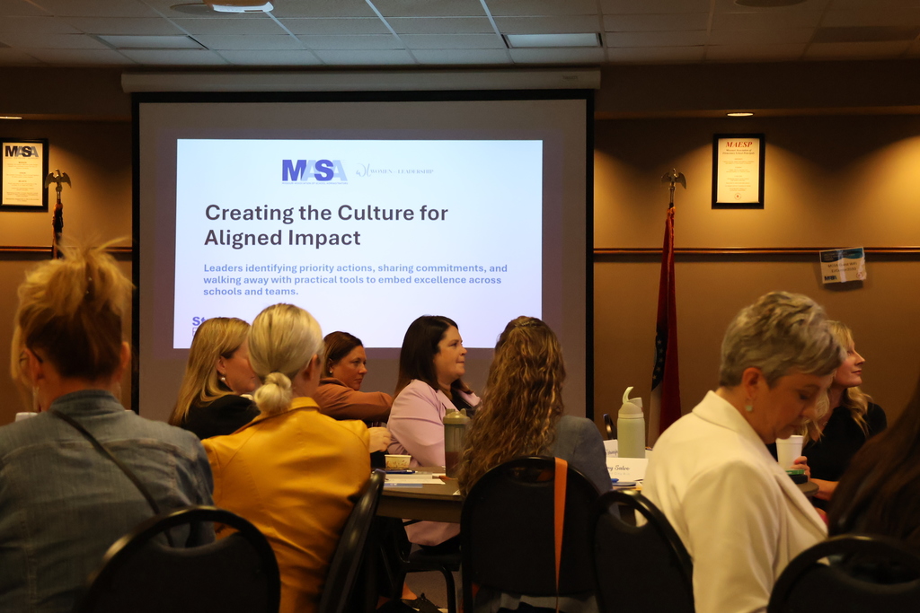 A room full of female leaders sitting at round tables. In the back a screen is displaying the presentation title "Creating the Culture for Aligned Impact."