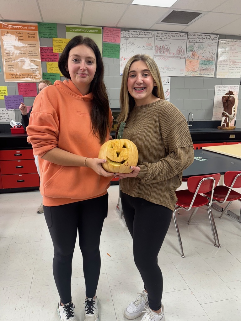 Two students smile while holding a small carved pumpkin with a triangle-eyed jack-o’-lantern face.