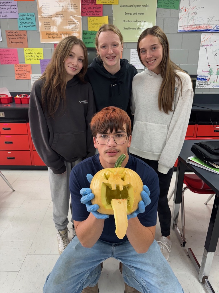 A group of five students pose together in a classroom. One student kneels in front holding a carved pumpkin with a square mouth and triangle eyes while wearing blue gloves. The others stand behind smiling.
