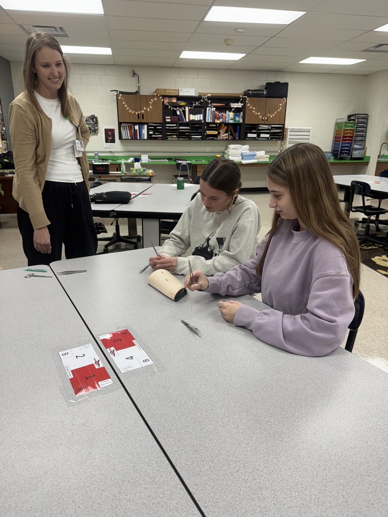 Two students sit at a classroom table practicing suturing techniques on a medical training pad as an instructor looks on and smiles. Surgical tools and instructional materials are on the table.