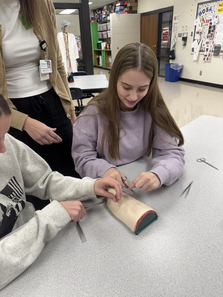 A student smiles while practicing suturing on a medical training pad as another student assists.