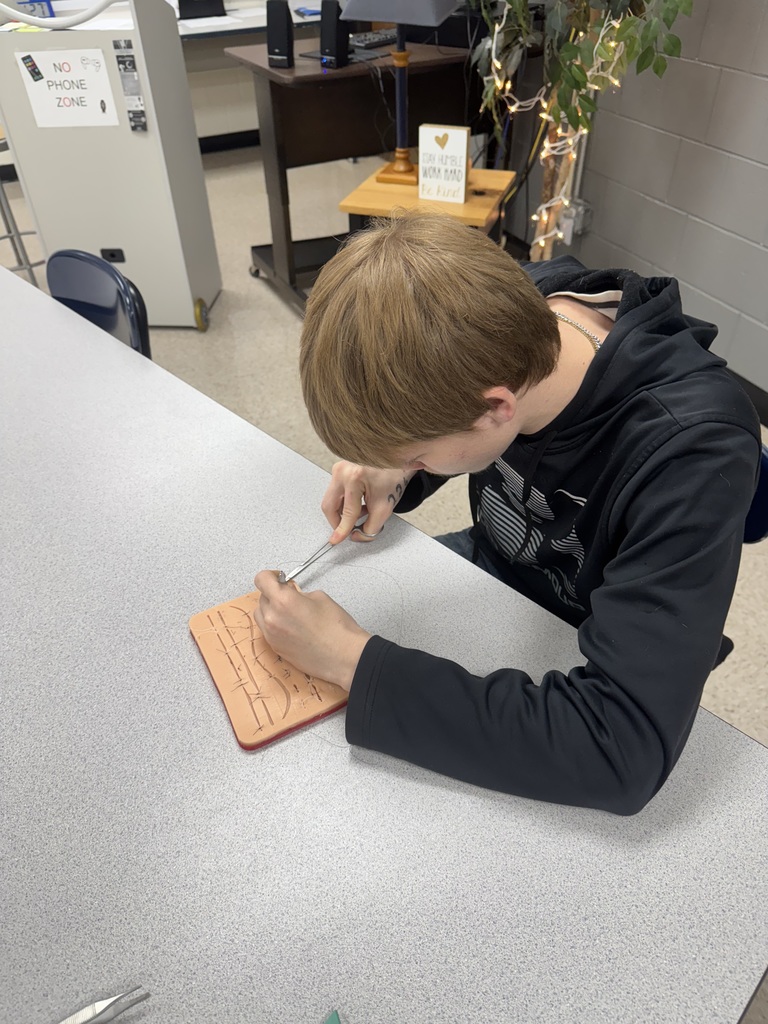 A student practices suturing on a silicone medical training pad at a classroom table.