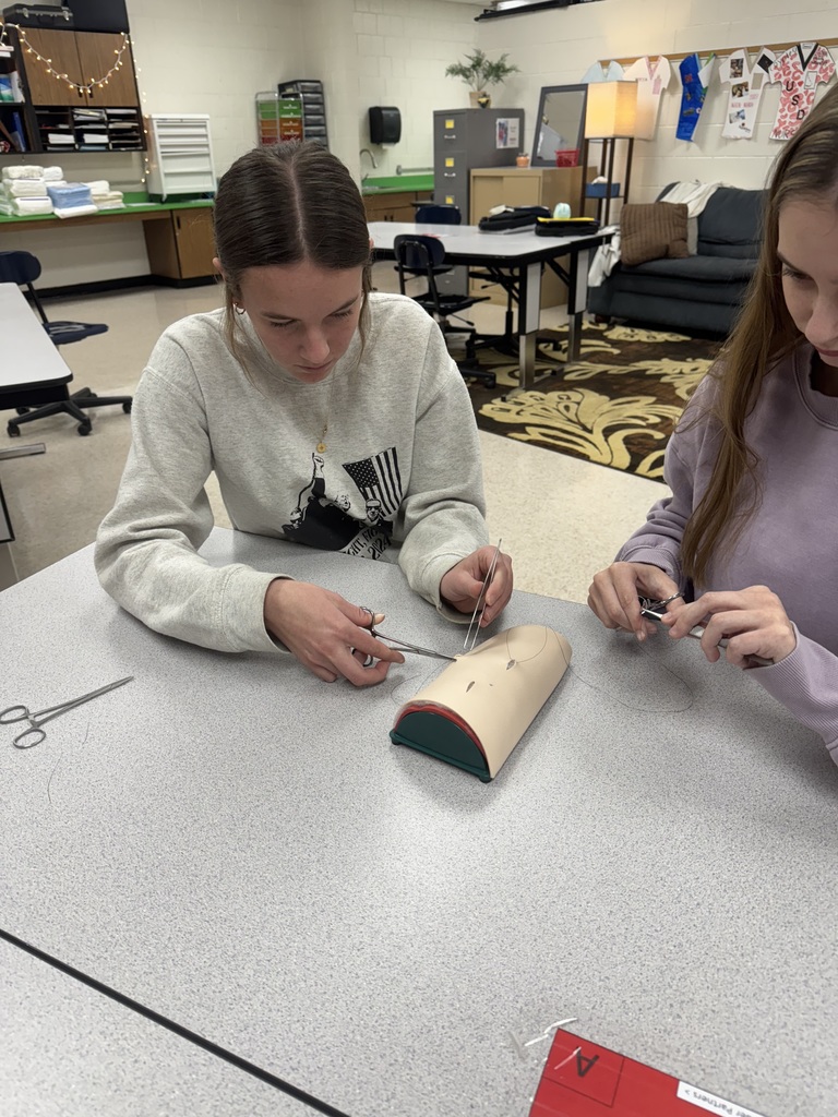 Close-up of two students using surgical instruments to practice suturing on a medical training pad. They are focused on their task while seated at a classroom table.