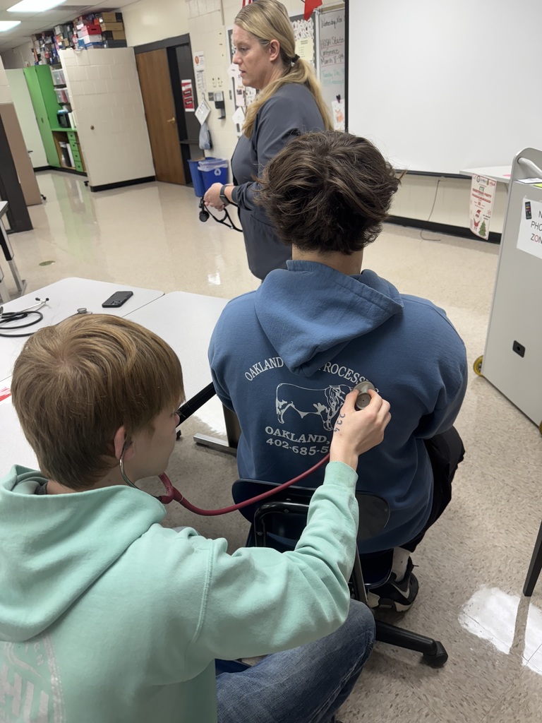 A student uses a stethoscope to listen to another student’s back while an instructor stands nearby demonstrating with her own stethoscope in a classroom.