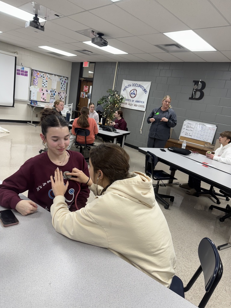A student listens to another student’s chest with a stethoscope at a classroom table while the instructor speaks to the class in the background.