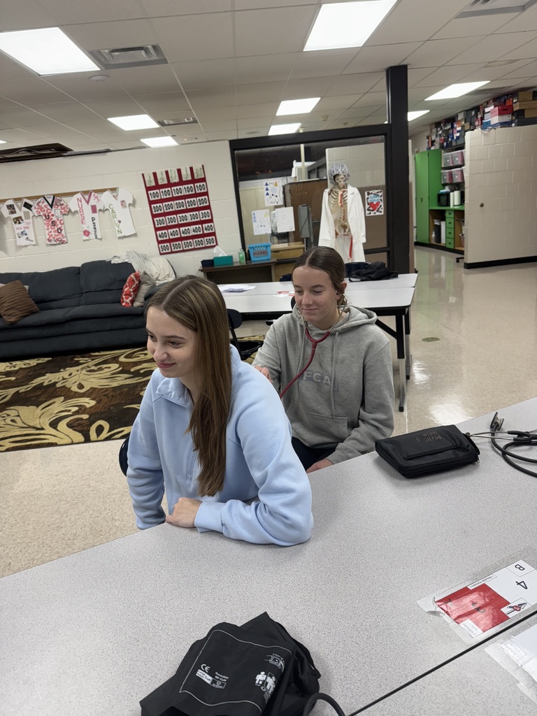 A student listens with a stethoscope to another student’s back while they sit at a classroom table.