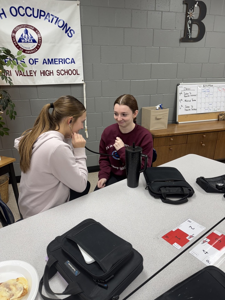 Two students sit facing each other as one uses a stethoscope to listen to the other’s chest in a classroom with HOSA materials visible on the wall.