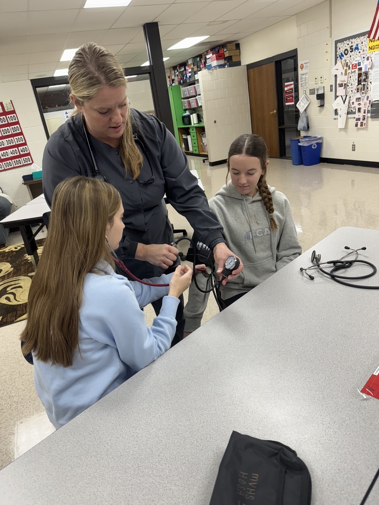 An instructor guides two students as they practice using a blood pressure cuff and stethoscope at a classroom table.