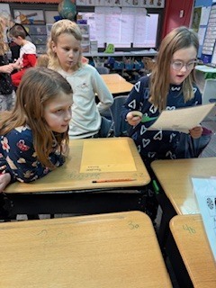 Three girls are grouped around desks in a classroom. One student stands and reads aloud from a paper while the others listen attentively.