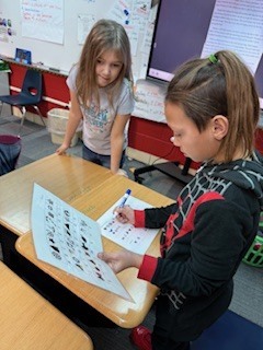 Two students work together at a desk. One girl writes on a worksheet while the other watches.