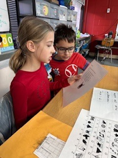 Two young students sit side by side at a classroom table, reading from a worksheet together. The girl in front holds the paper while the boy leans in to follow along.