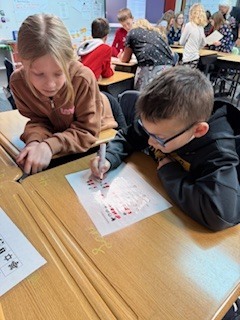 A boy and girl work on a red-marked worksheet together at a wooden desk. The boy writes with a marker while the girl observes.