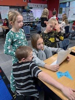Four elementary students gather around a desk, looking at a shared sheet of paper. Two students sit while two others stand, all focused on the activity.