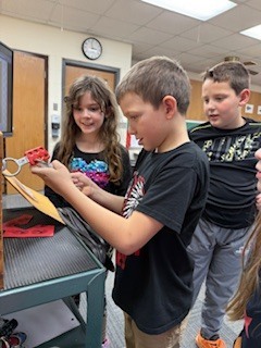 Three students work on a lockbox task at a cart. One boy is focused on opening a red lock while two others watch with interest.