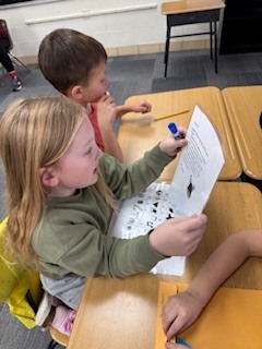 A girl sitting at a desk reads and marks answers on a worksheet while a classmate beside her participates.