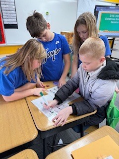 Four students collaborate at a desk, solving a worksheet with green and purple markers.