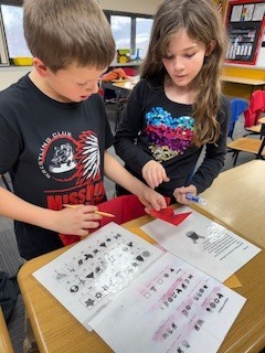Two students work at a desk on a decoding activity using a worksheet and red filter.