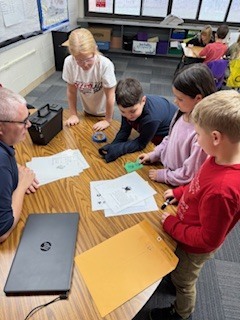 A group of students and an adult gather around a table looking at printed worksheets and activity papers.