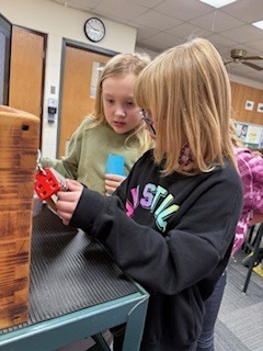 Two girls examine a red lock on a wooden lockbox cart, working together to figure out the combination.