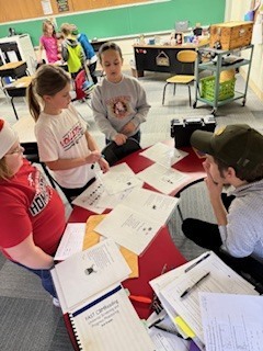 Five students stand around a red and white table covered with worksheets, engaged in a group decoding or problem-solving activity.