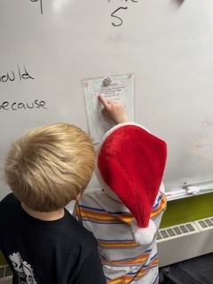 Two boys look at a printed clue posted on a whiteboard. One of them, wearing a red Santa hat, is pointing at the text while the other observes.