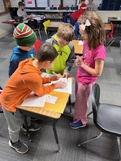 Four students work together at a desk, closely examining papers and what appears to be a puzzle or worksheet. One student points while others observe and discuss.