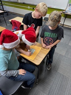 A group of four boys sits around a desk, working on a paper clue. Two students wear red Santa hats.