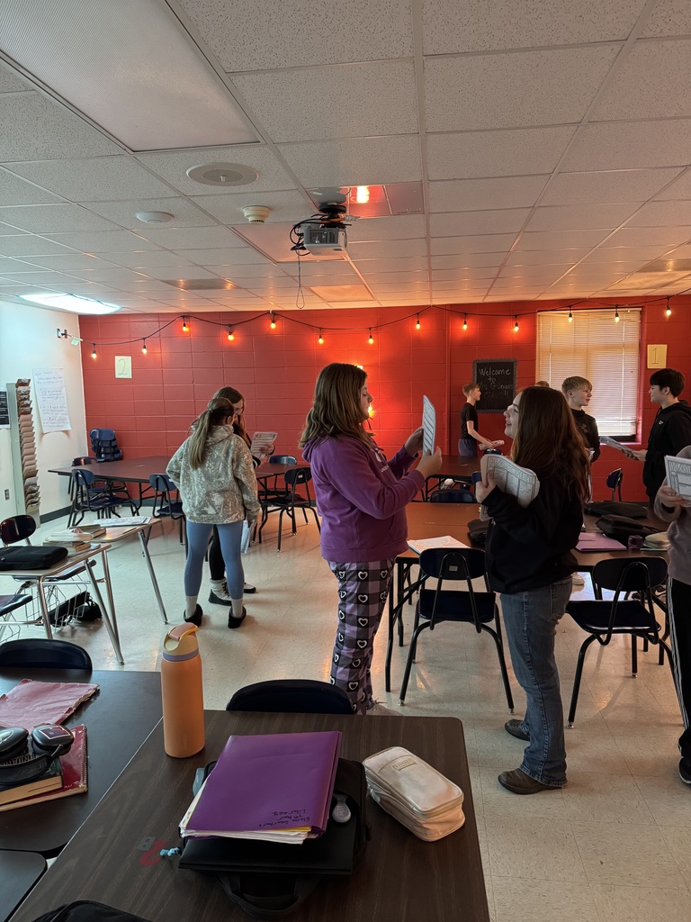 Students stand in pairs around a classroom with orange lighting, reading and discussing worksheets.