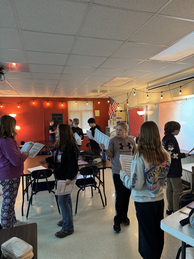 Students actively engage in a partner reading activity, holding worksheets and moving around the classroom decorated with string lights.