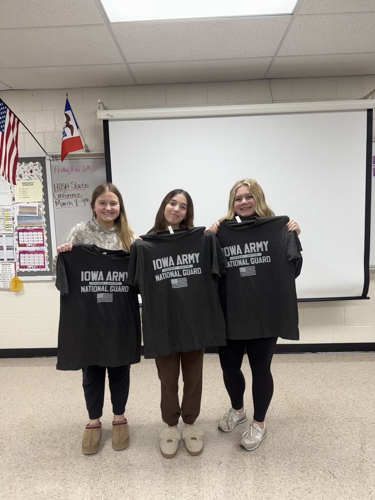 Three students smile while holding up Iowa Army National Guard t-shirts in front of a whiteboard.