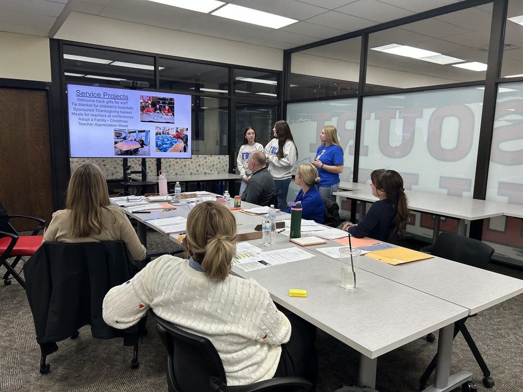 Students present service projects to a group of seated adults in a meeting room.