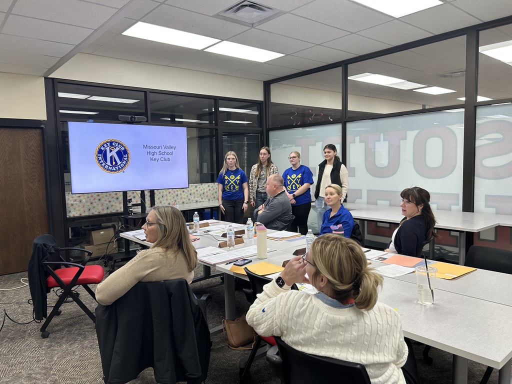 High school Key Club members stand and present to a seated audience in a meeting room.