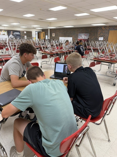 Three students collaborate around a laptop while reviewing materials during class group work.