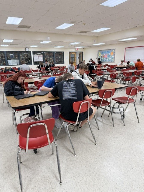 Multiple student groups work independently at cafeteria tables using laptops and notes during class.