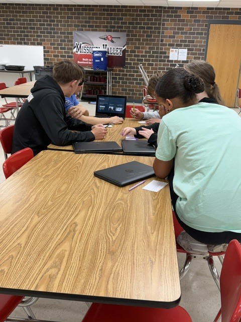 Students work together at cafeteria tables using laptops and notes during a collaborative classroom activity.