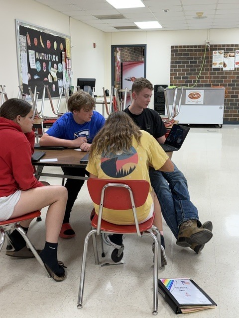 Multiple student groups work independently at cafeteria tables using laptops and notes during class.