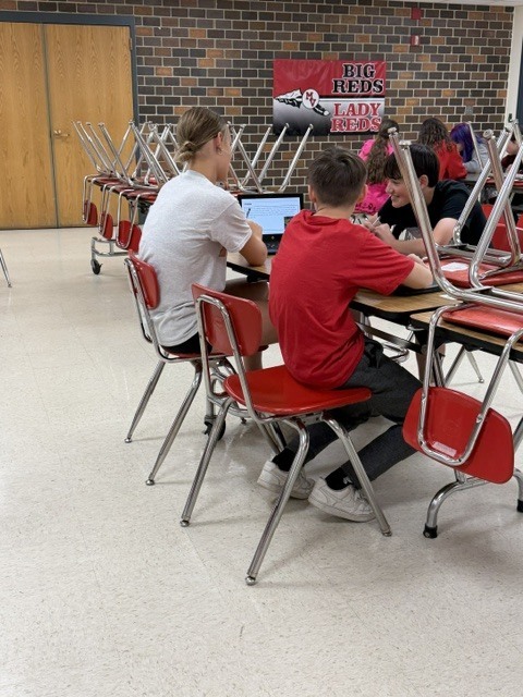 Three students sit together using a laptop and discussing assignments at a cafeteria table.