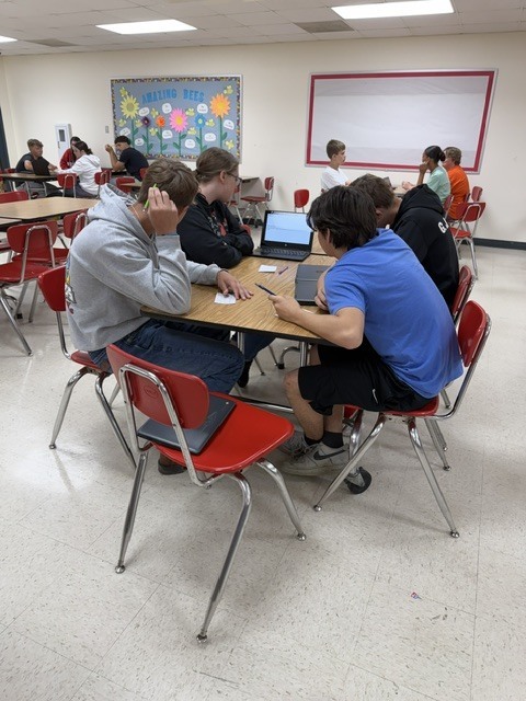 Several student groups collaborate at tables with laptops and papers during a class activity in the cafeteria.