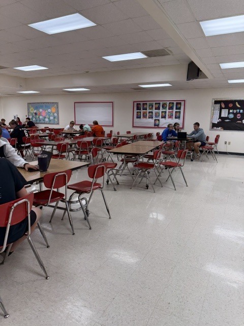 Students spread throughout the cafeteria working in small groups with laptops and classroom materials.