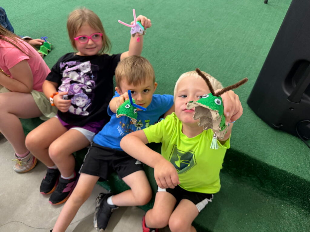 Kids sitting on a green rug holding up frog crafts